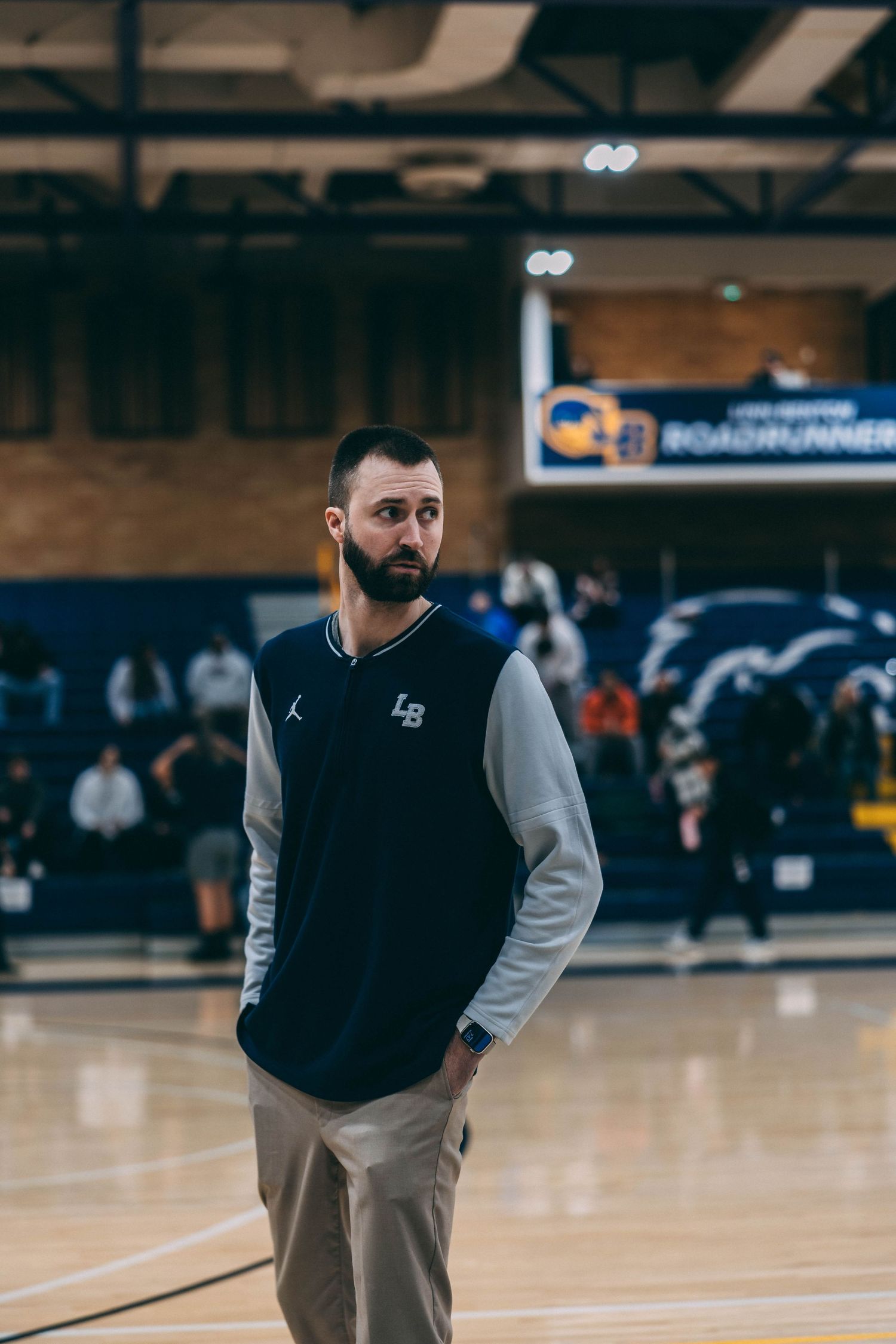 Coach in black and gray vest watches basketball practice in indoor gymnasium.