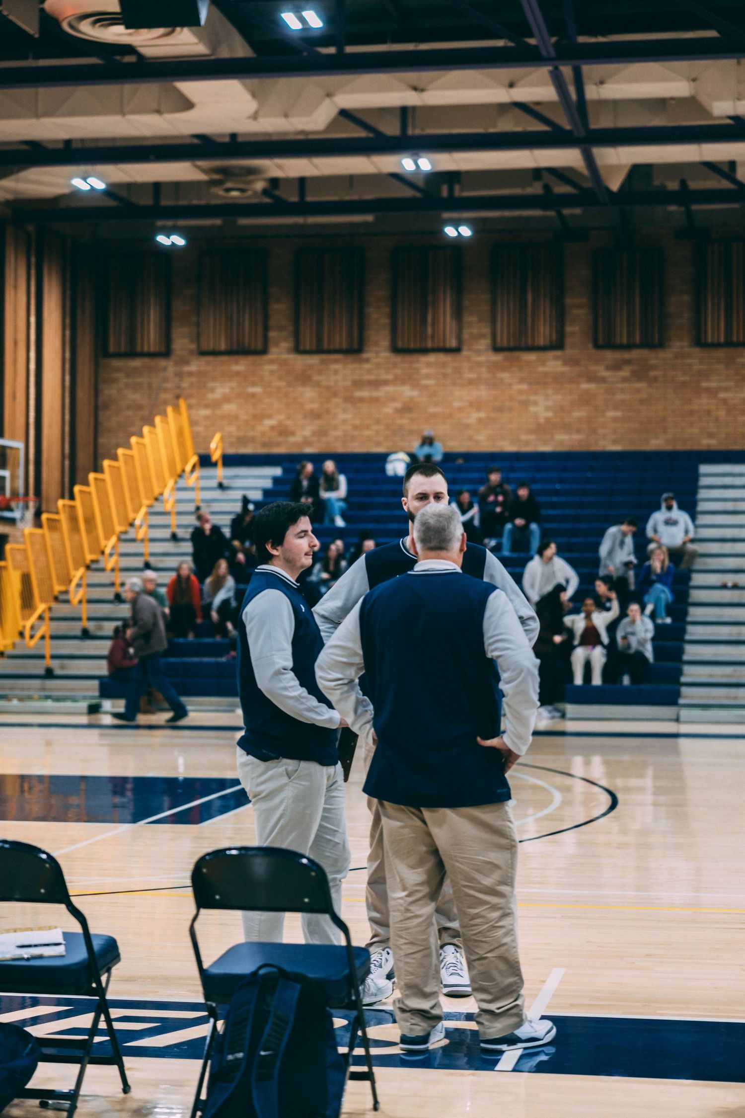Basketball officials and coaches gather on the sidelines of an indoor gymnasium court during a game.