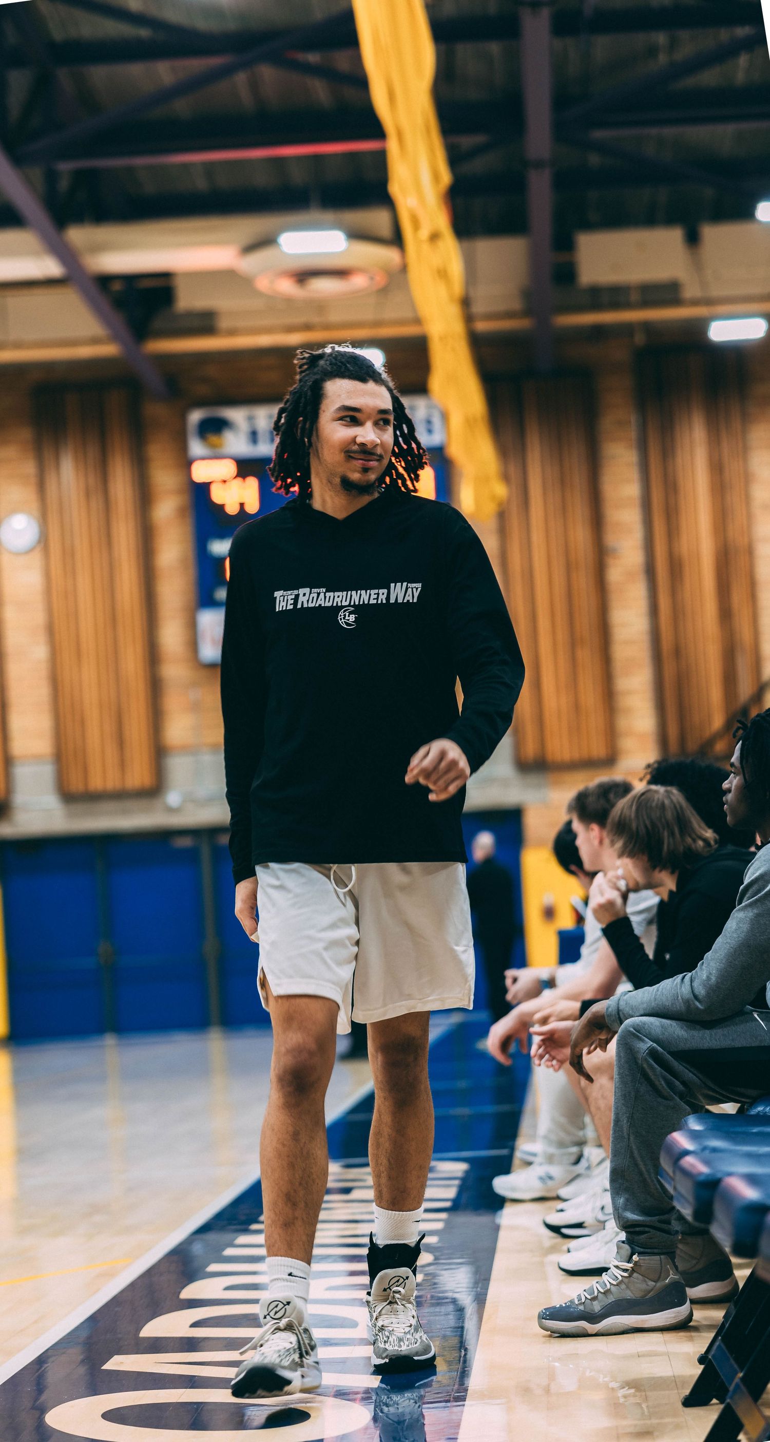 Player in black hoodie stands on the sideline of an indoor basketball court.