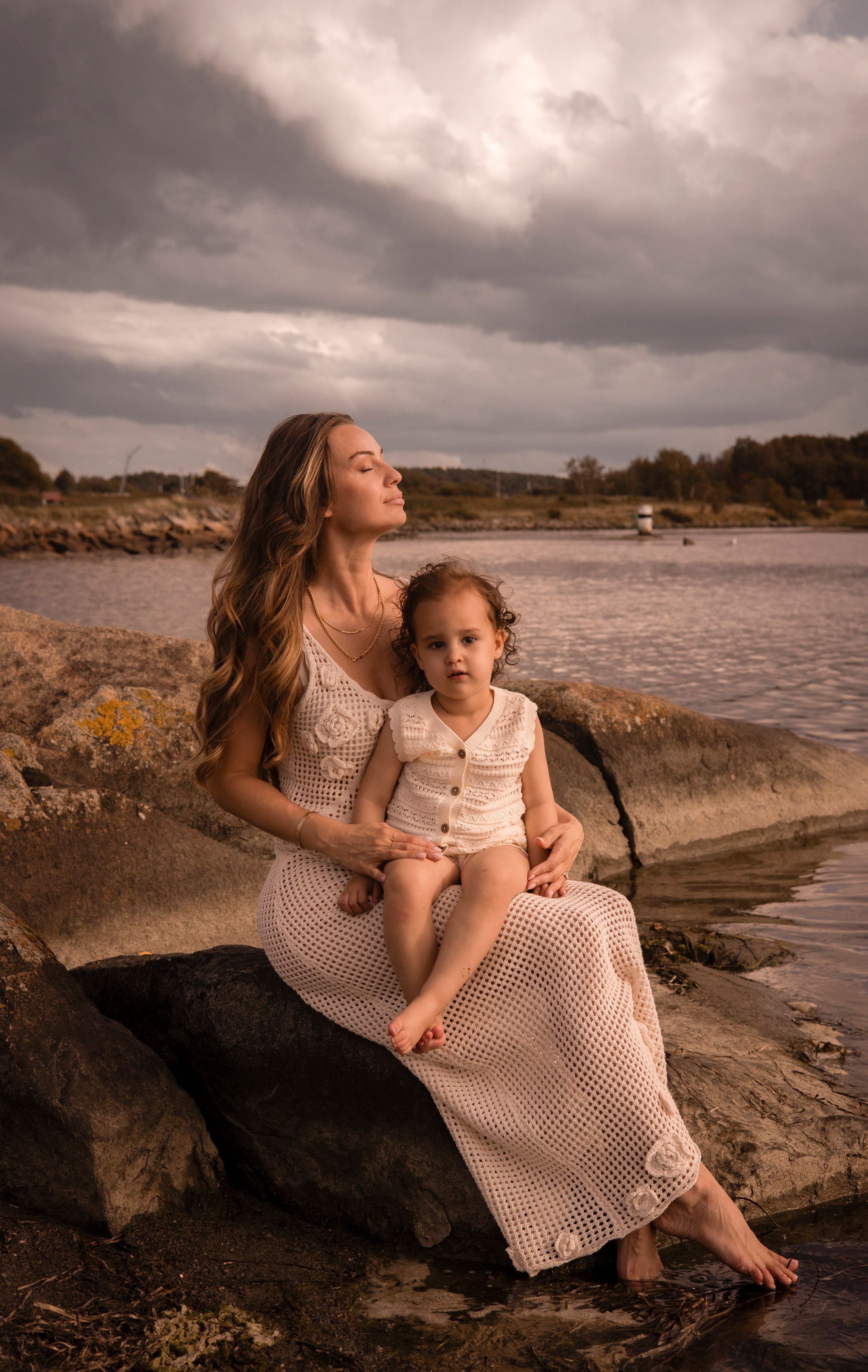 mamma sitter på en klippa med sitt barn i sin famn. hon blundar med ansiktet ut mot havet. barnet kollar in i kameran.