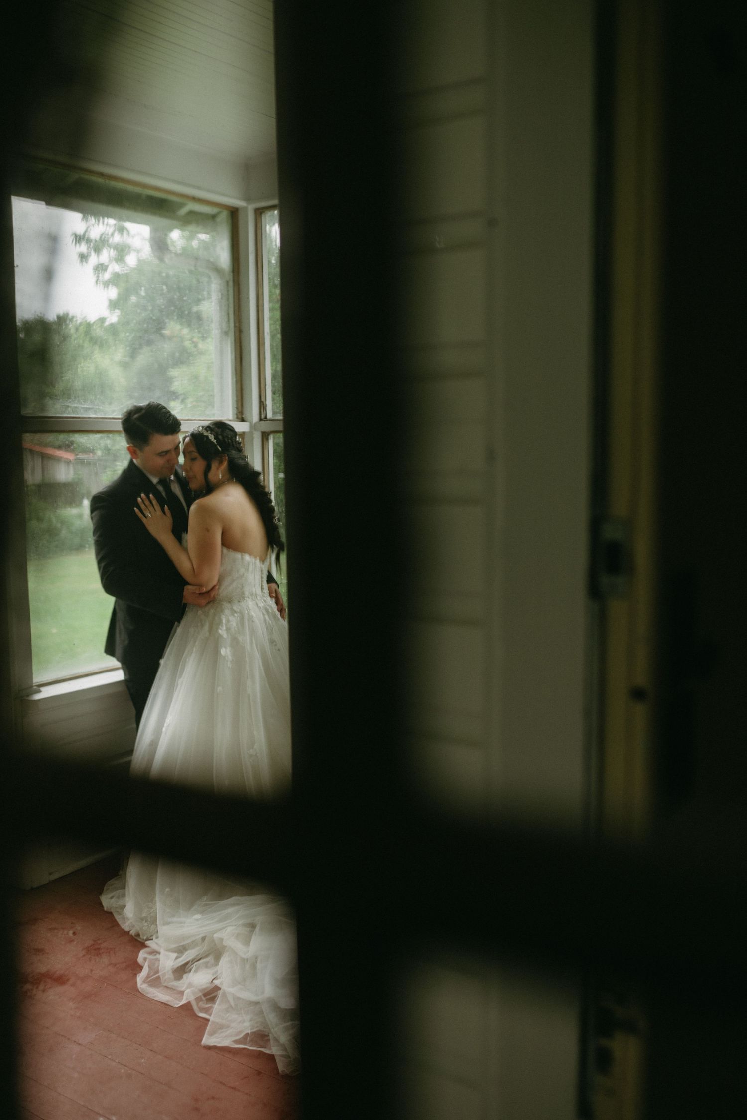 A romantic wedding couple shares an intimate moment by a window in a dimly lit room.
