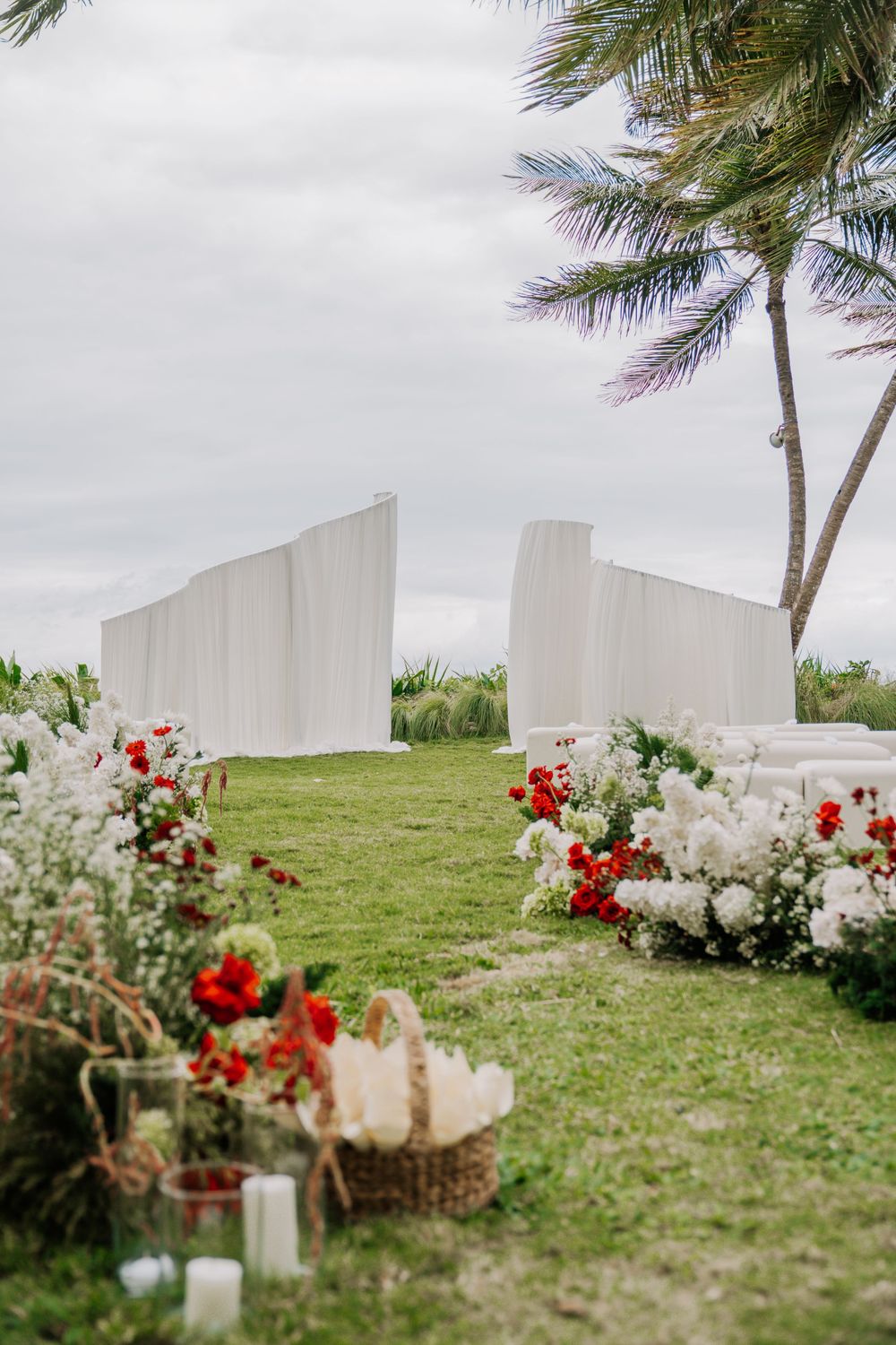 White modern wedding ceremony backdrop with red and white floral arrangements and palm trees against cloudy sky.