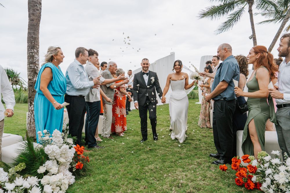 Wedding guests gather outdoors near palm trees and ocean for a tropical beach ceremony celebration.