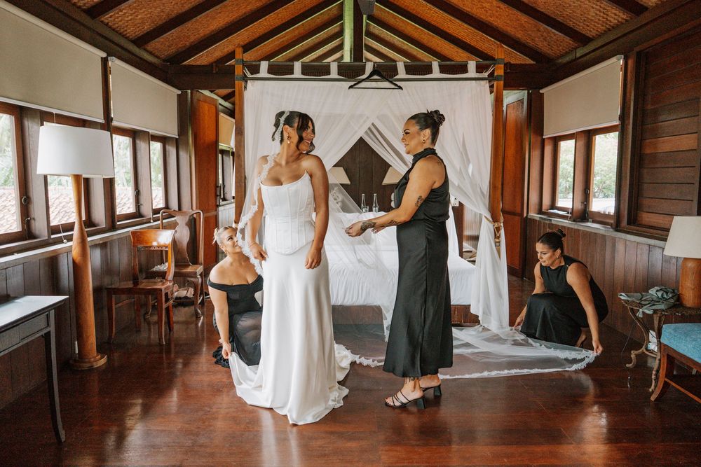Bridesmaids in sage green dresses help bride get ready in rustic wooden cabin before wedding ceremony.