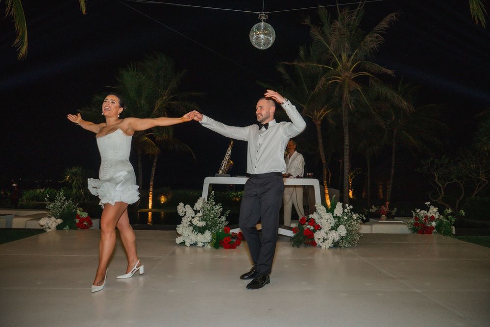 A couple performs a playful dance move during a nighttime outdoor wedding reception with palm trees in the background.