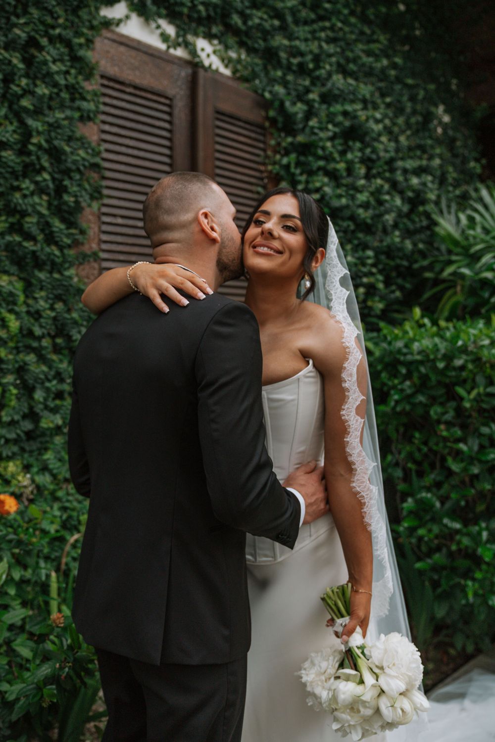 Romantic wedding couple embraces in a garden setting with lush green foliage and white flowers.