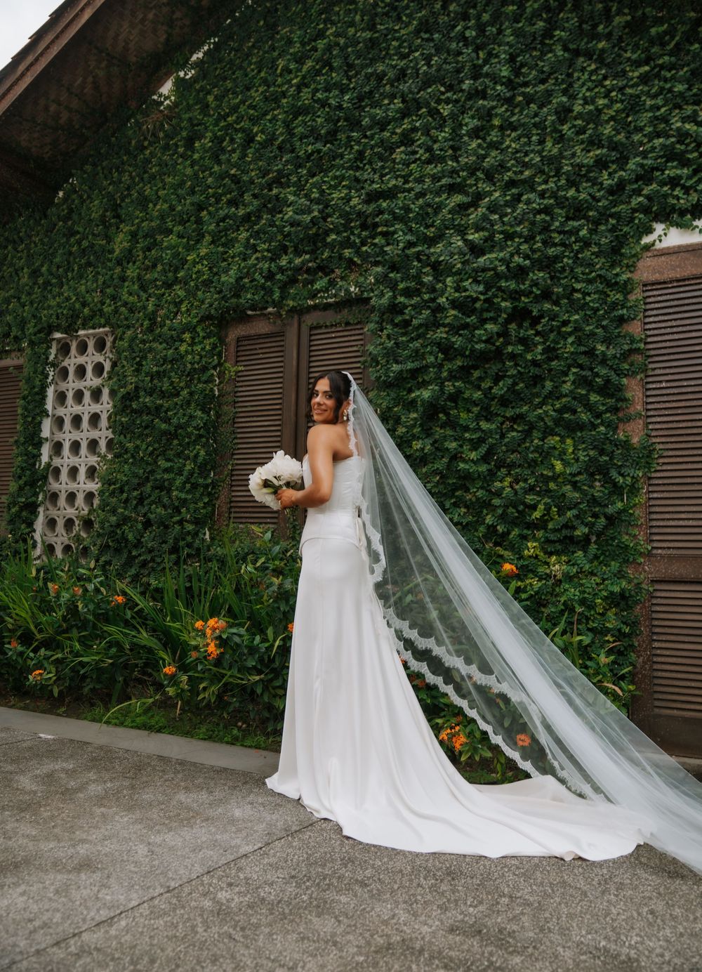 A bride in a white wedding gown and veil poses against a lush green ivy-covered wall with yellow flowers.