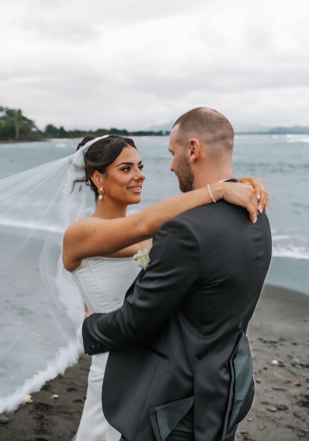 A couple shares a romantic moment on a cloudy beach, with the bride's veil flowing in the ocean breeze.