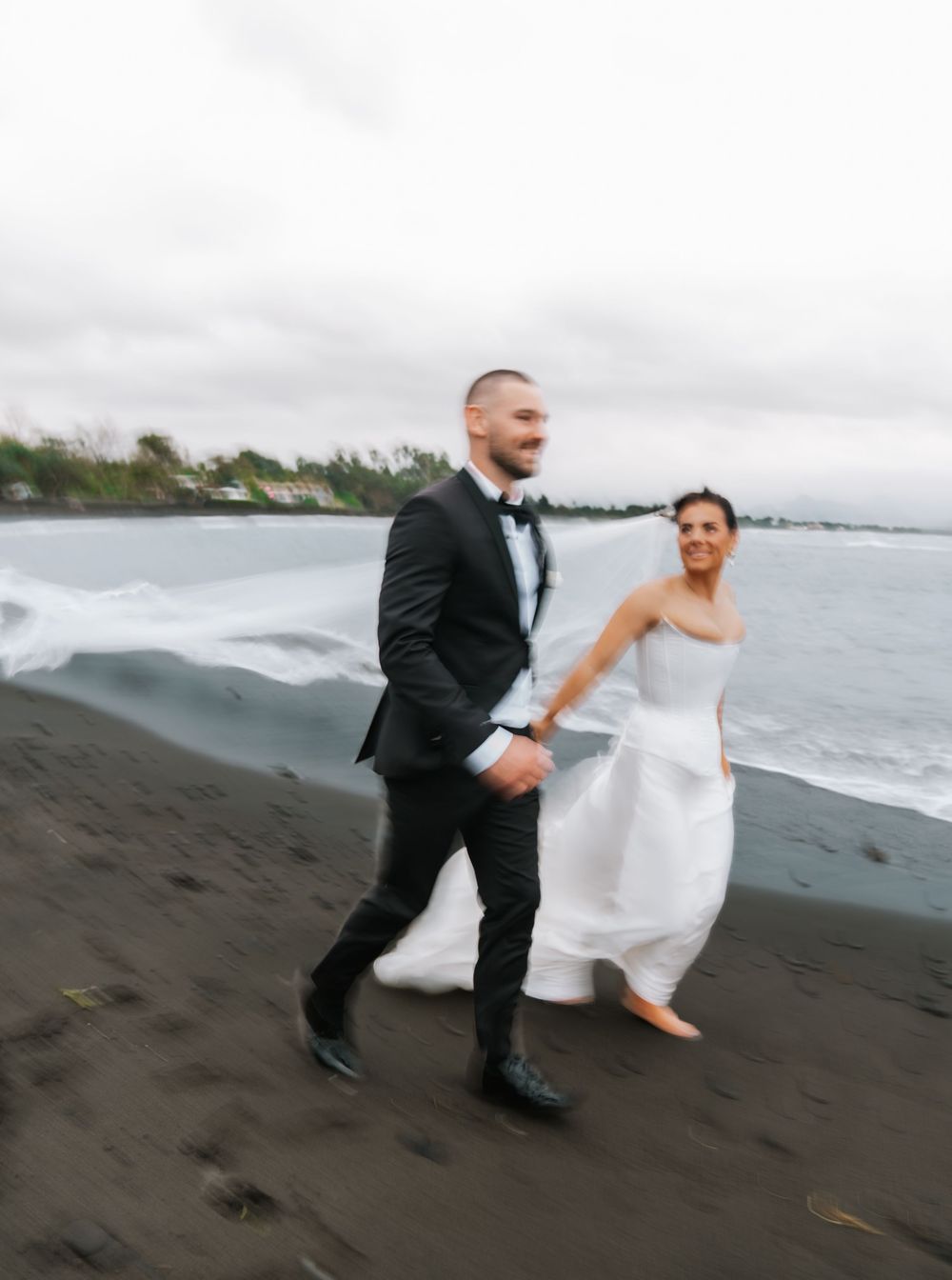 A couple in wedding attire runs along a black sand beach with ocean waves and cloudy skies in the background.