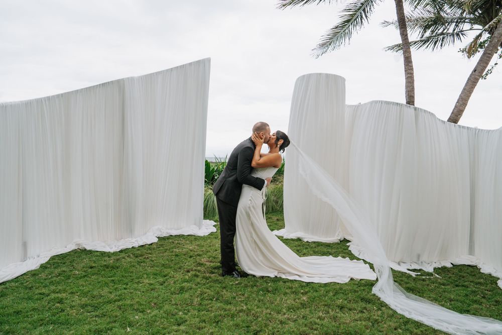 A romantic wedding couple poses with flowing white fabric in a lush tropical garden setting.