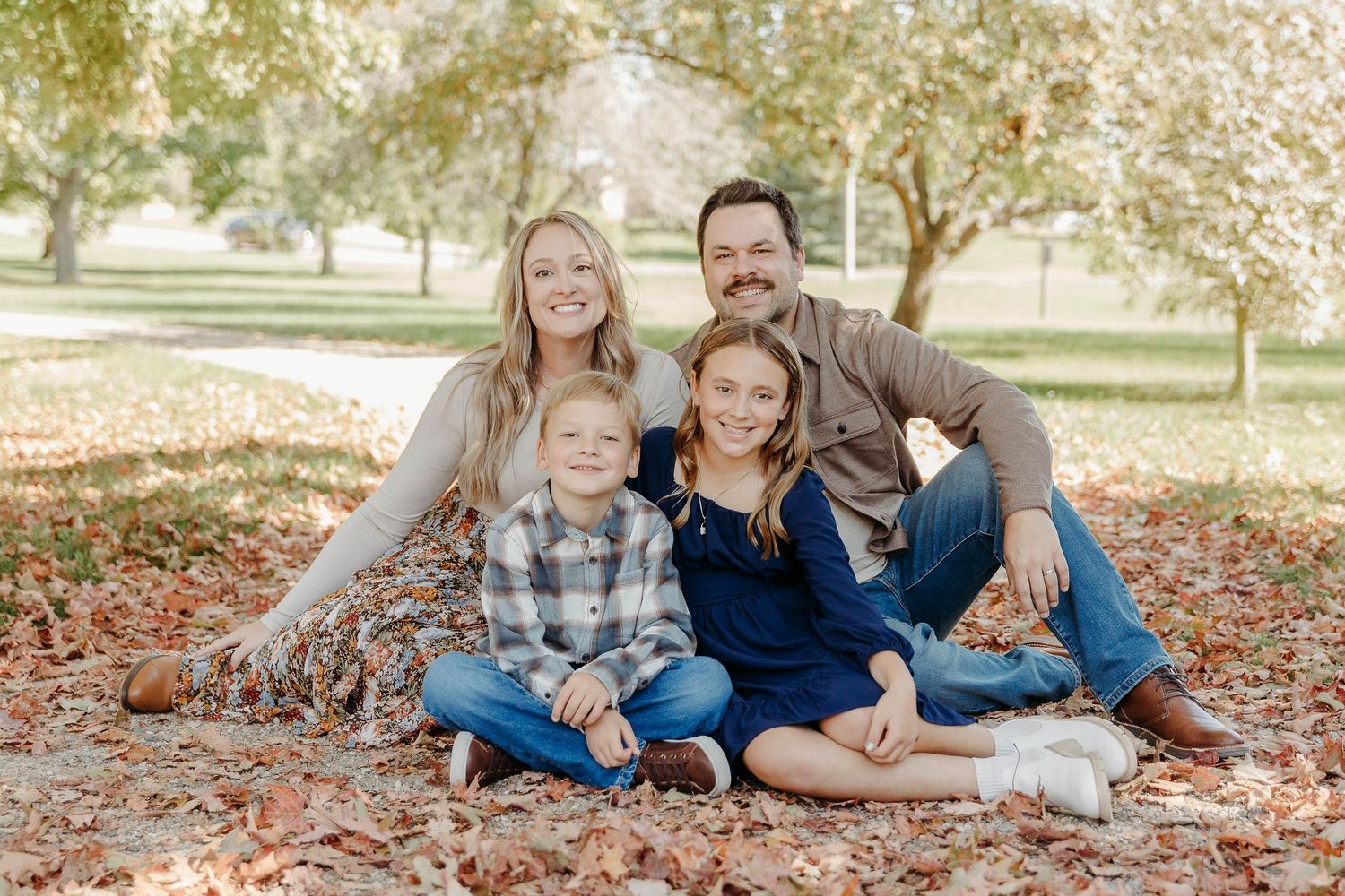 A family sits together on fallen autumn leaves during an outdoor photo session in a park setting.