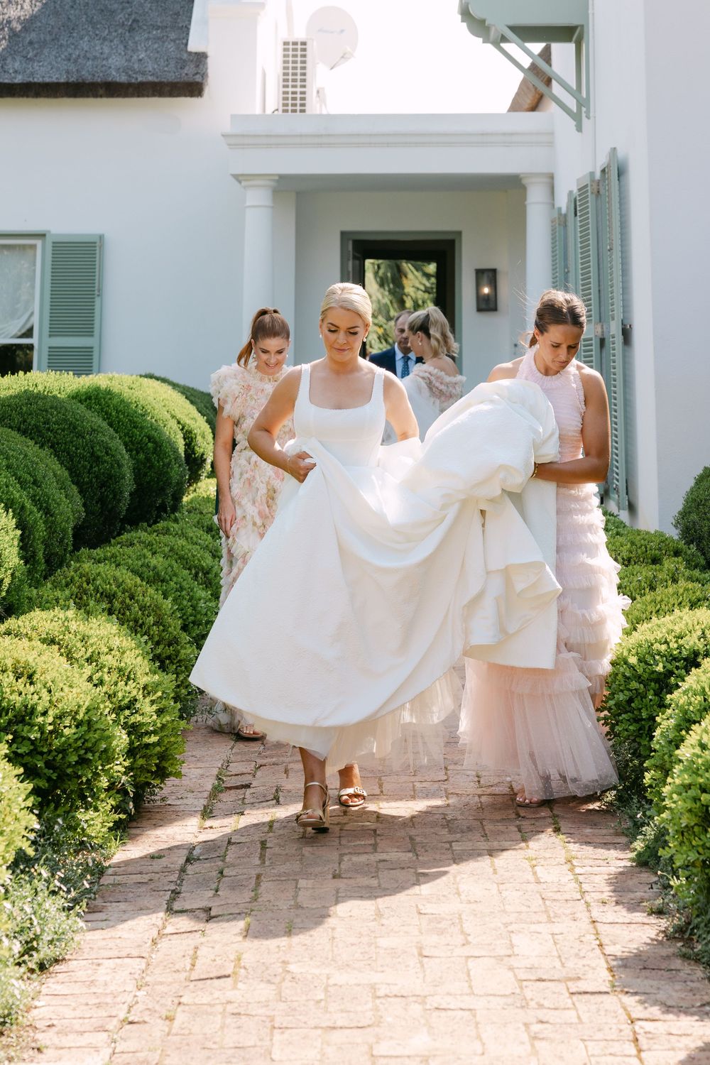 Bridesmaids in blush dresses help adjust the bride's flowing white wedding gown on a brick patio.