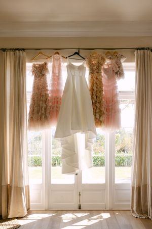 Wedding dresses hanging in front of sunlit windows with flowing curtains.