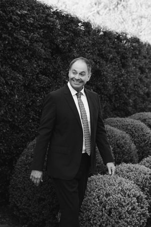 Black and white sequence of a person in a suit walking and smiling near a garden hedge.