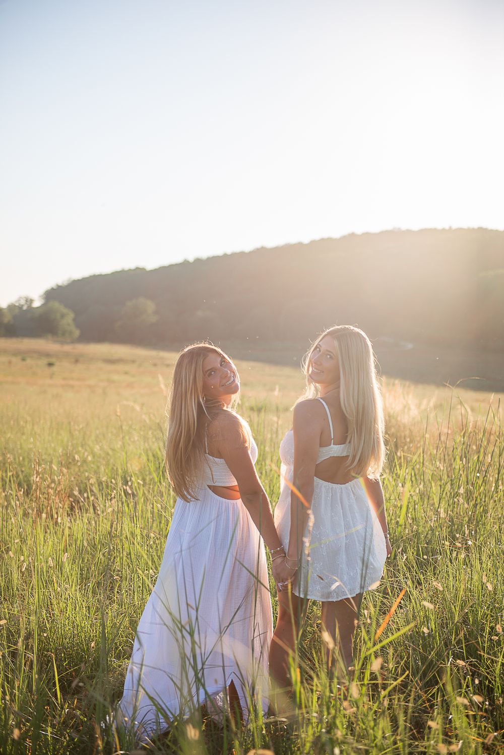 Two people in white dresses walk together through a sunlit meadow of tall grass at golden hour.