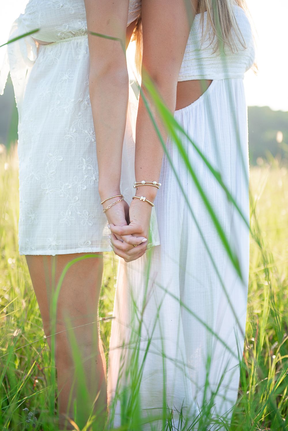 Two people holding hands in white dresses stand among tall grass at sunset.