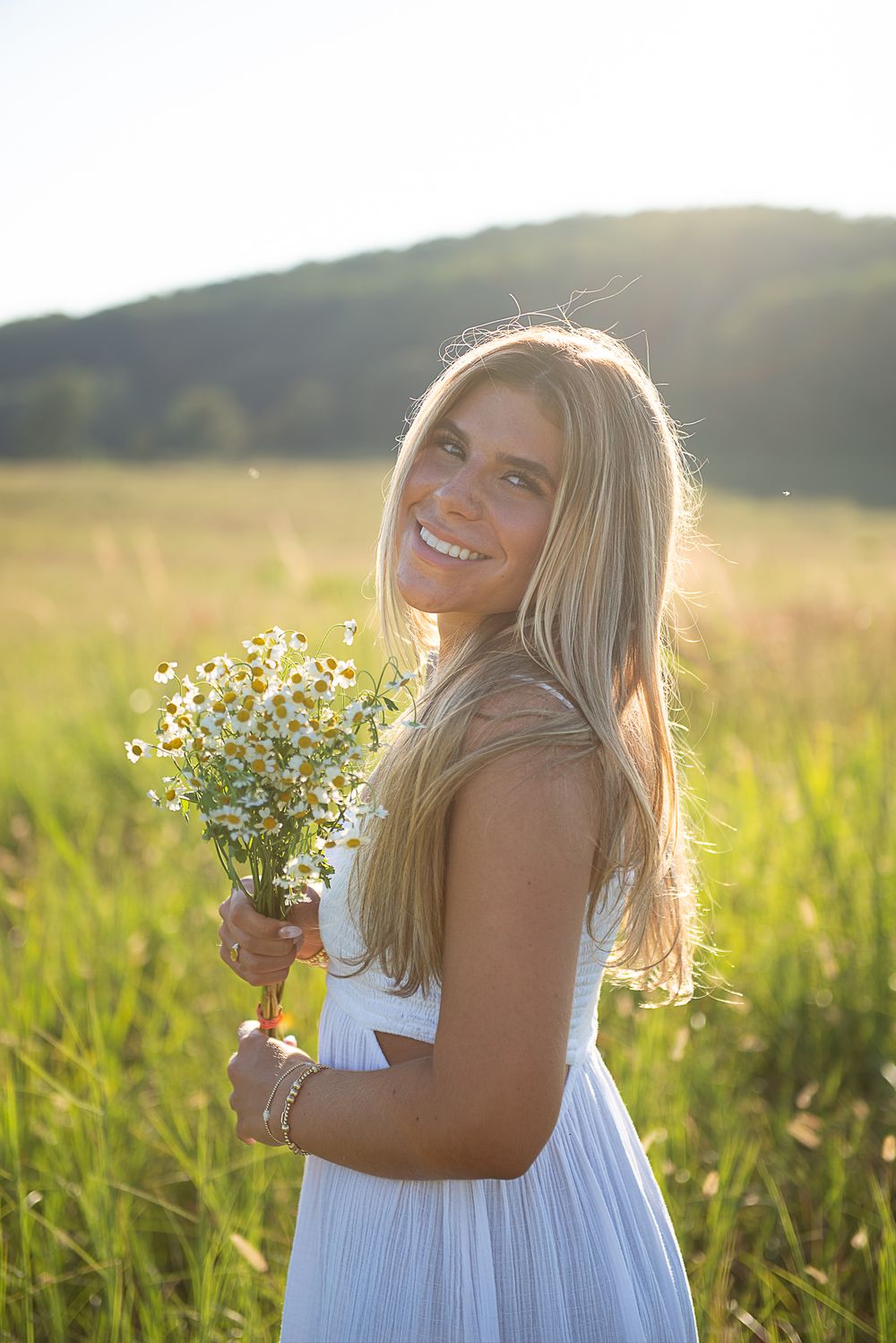 A woman in a white dress holds wildflowers while standing in a golden summer meadow.