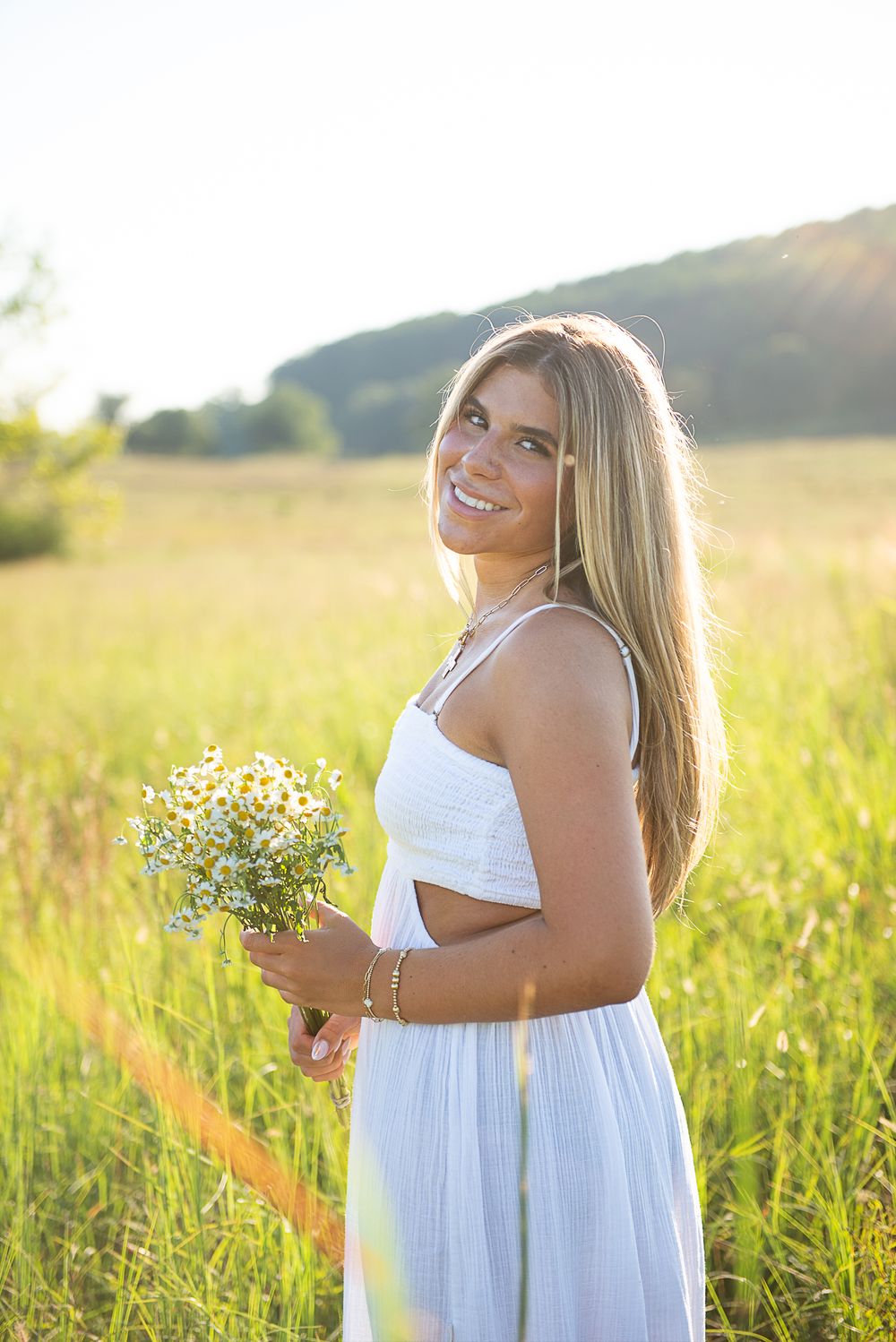 A woman in a white dress holds wildflowers while standing in a sunlit meadow during golden hour.