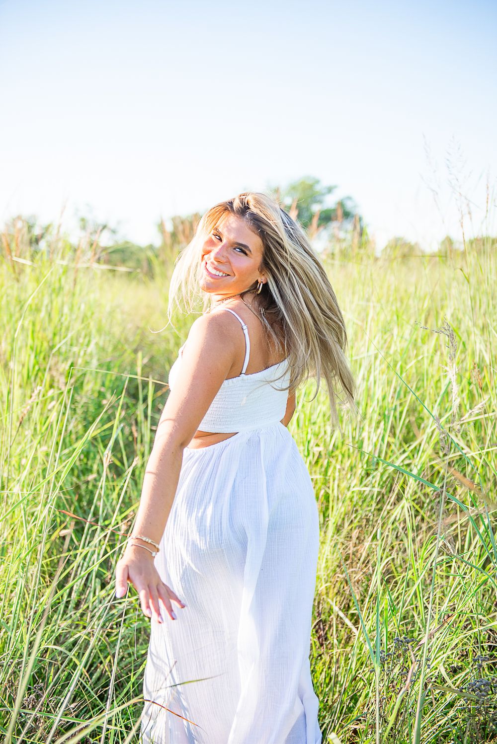 Woman in flowing white dress twirls joyfully in tall summer grass field at golden hour.