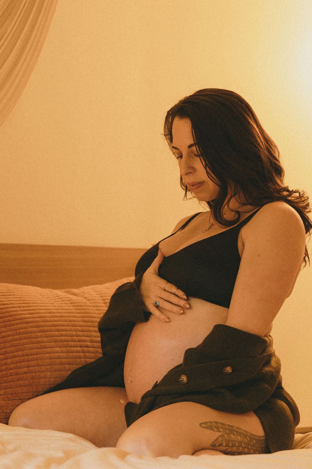 Smiling pregnant woman in black bralette and jacket tenderly holding her bare baby bump on a bed.
