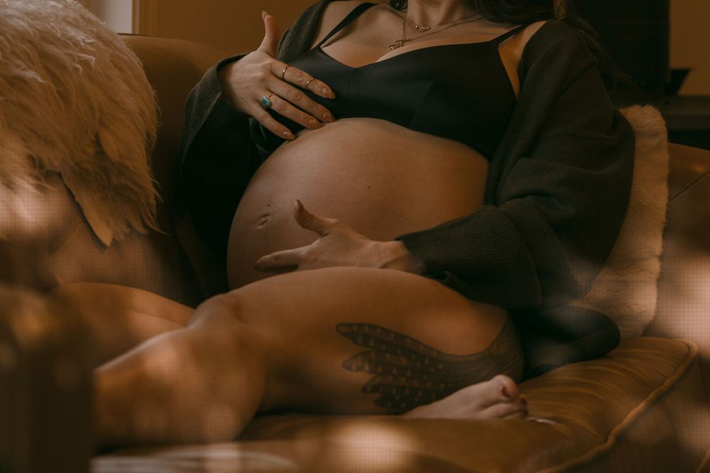 Close-up of pregnant woman in black lingerie gently holding her bare baby bump with tattooed thigh.