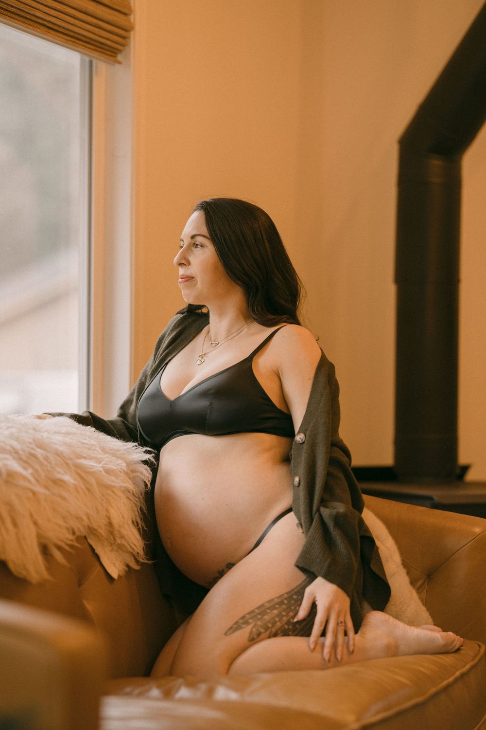Pregnant woman in black bralette and jacket sitting on leather sofa gazing out a sunlit window.