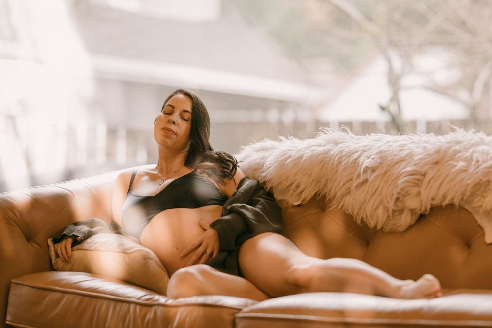 Pregnant woman in black lingerie reclining on a leather sofa with fur throw in natural light.