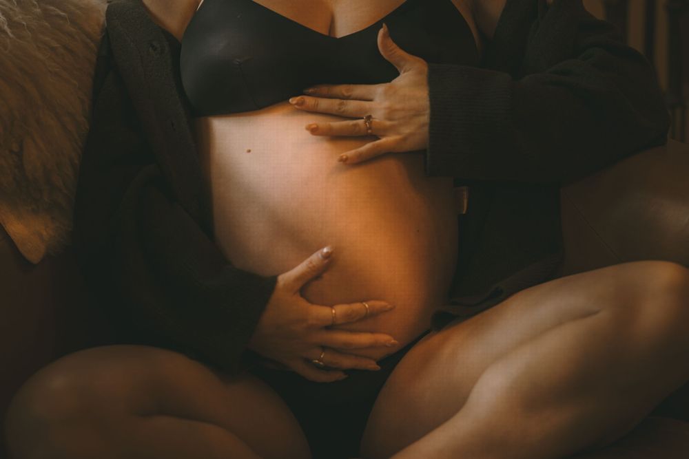 Close-up of pregnant belly being cradled by hands wearing black lingerie and open jacket.