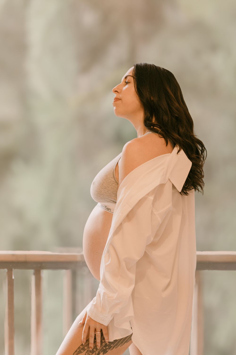Pregnant woman in beige bralette and open white shirt standing on balcony with green backdrop.