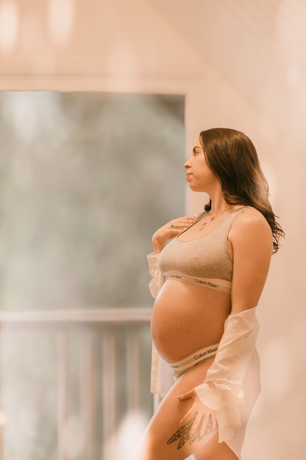 Pregnant woman in Calvin Klein underwear and open white shirt standing by large window indoors.