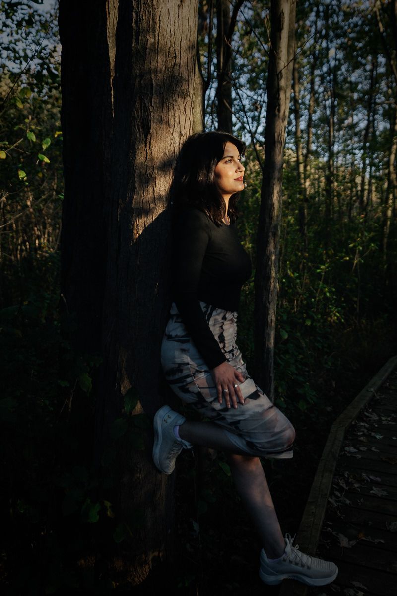 A figure sits against a tree trunk in a moody forest setting with dramatic lighting and shadows.