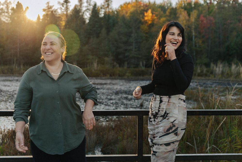 Two people share a joyful moment while leaning against a railing at sunset with autumn trees in the background.