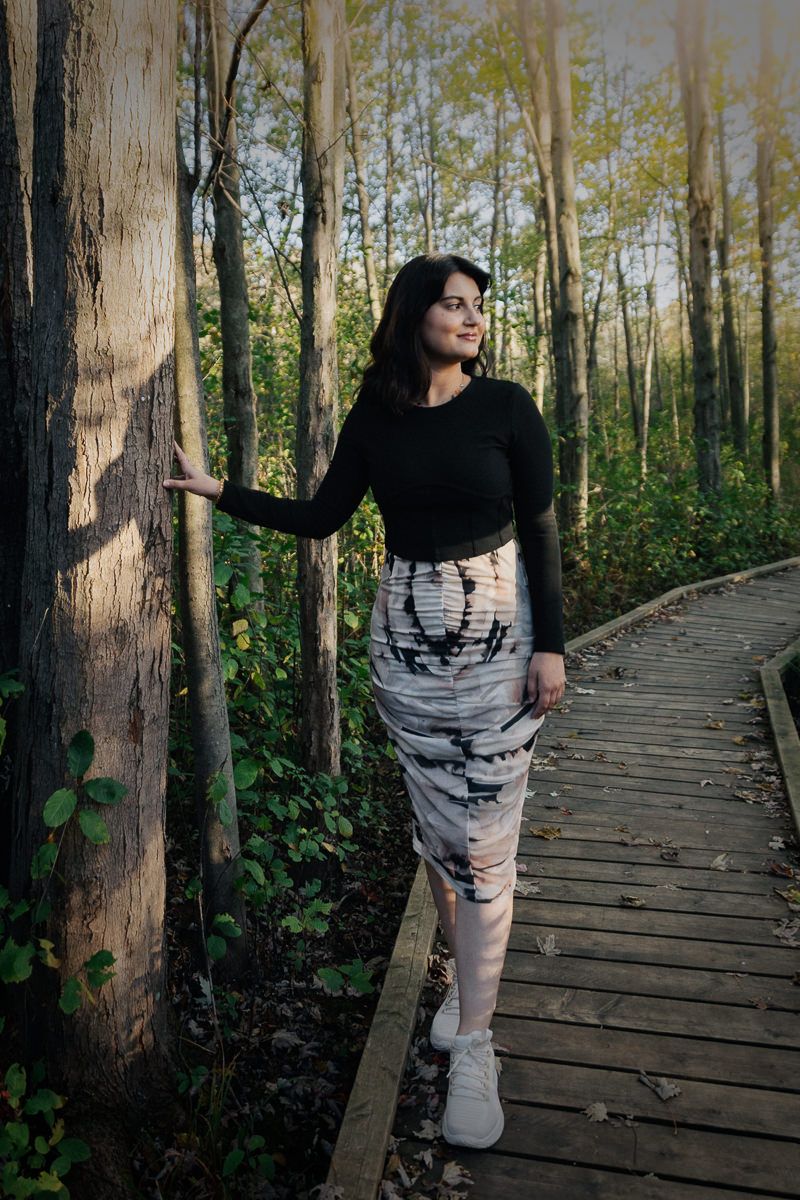 A person in black and gray clothing poses on a wooden boardwalk path through a sunlit forest.