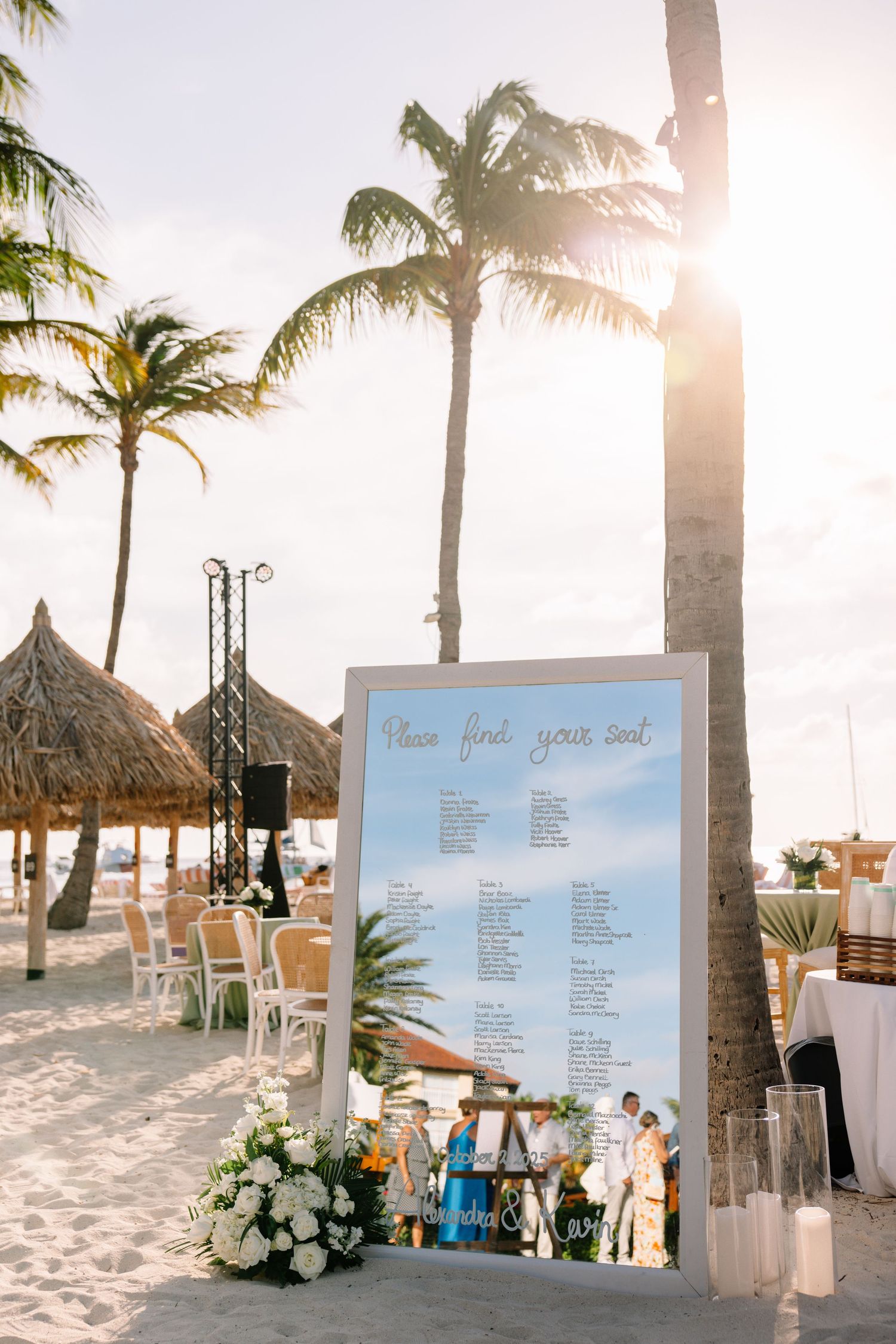 Beach wedding seating chart display with palm trees and tiki huts at sunset.