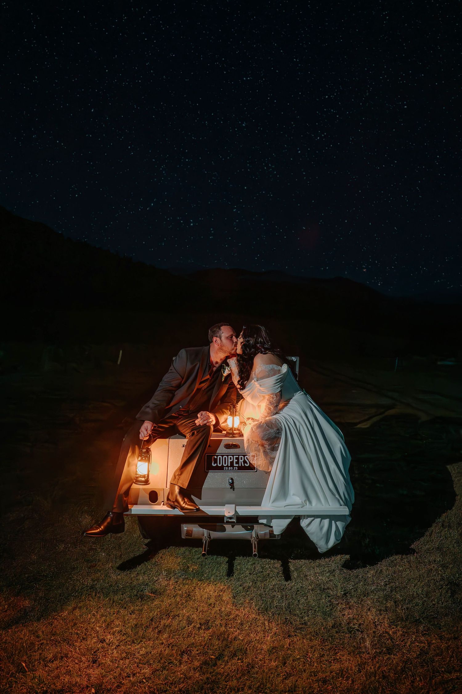 A couple shares a romantic moment at a picnic table under a starry night sky with warm lantern lighting.
