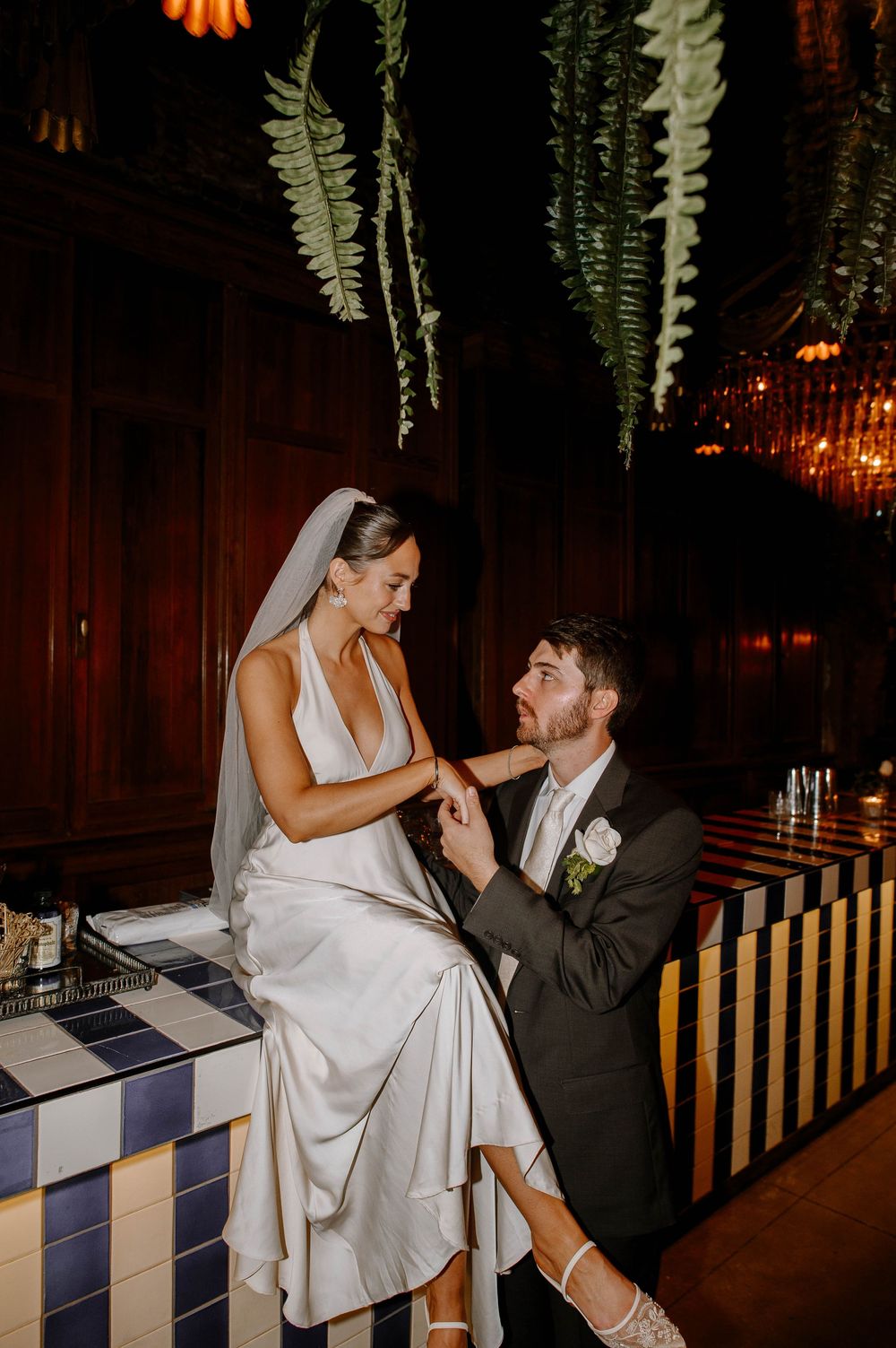 A couple shares a playful moment on a striped bench under hanging ferns at an evening wedding reception.