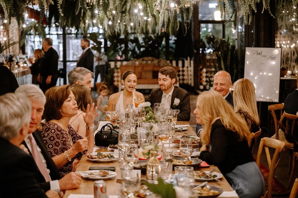 Elegant dinner party guests seated at a long table with wine glasses and floral centerpieces in a warmly lit restaurant.