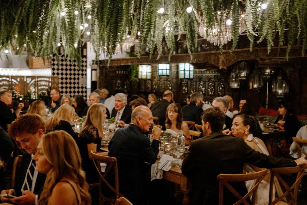Wedding reception guests gathered in an elegant venue under hanging floral decorations and string lights.