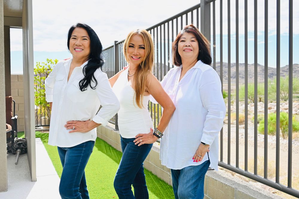 Three stylish women wearing white tops and blue jeans smile together on a green turf patio with mountain views.