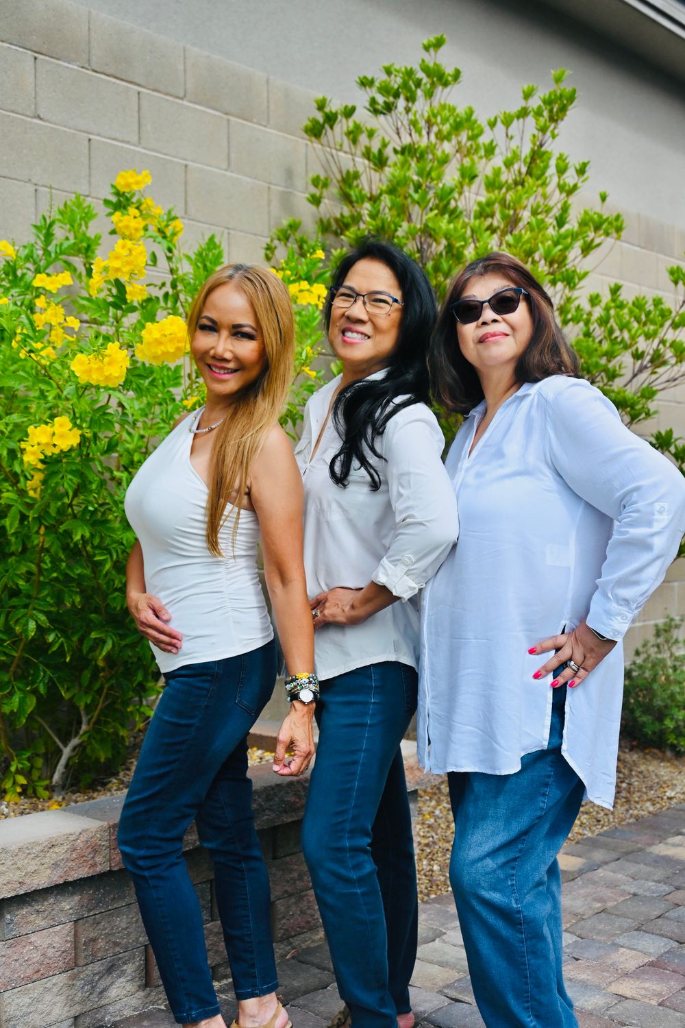 Three women in white tops and jeans posing outdoors near yellow flowering bushes.