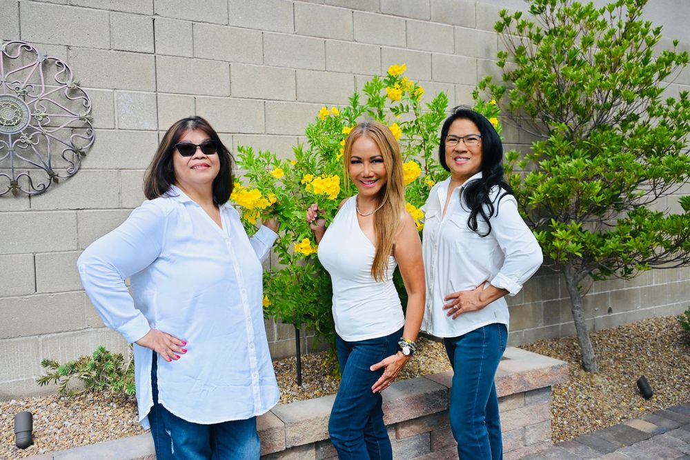 Three women in white shirts and jeans posing together in a sunny outdoor garden.