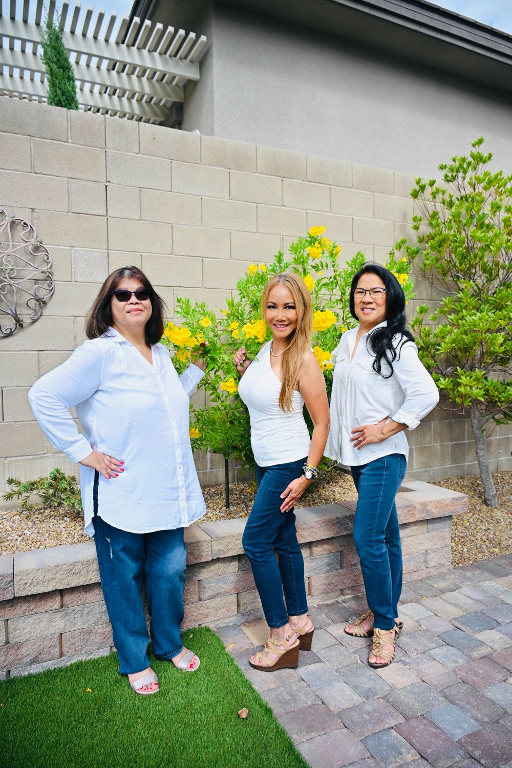 Three women in white tops and jeans posing in a backyard with yellow flowers.