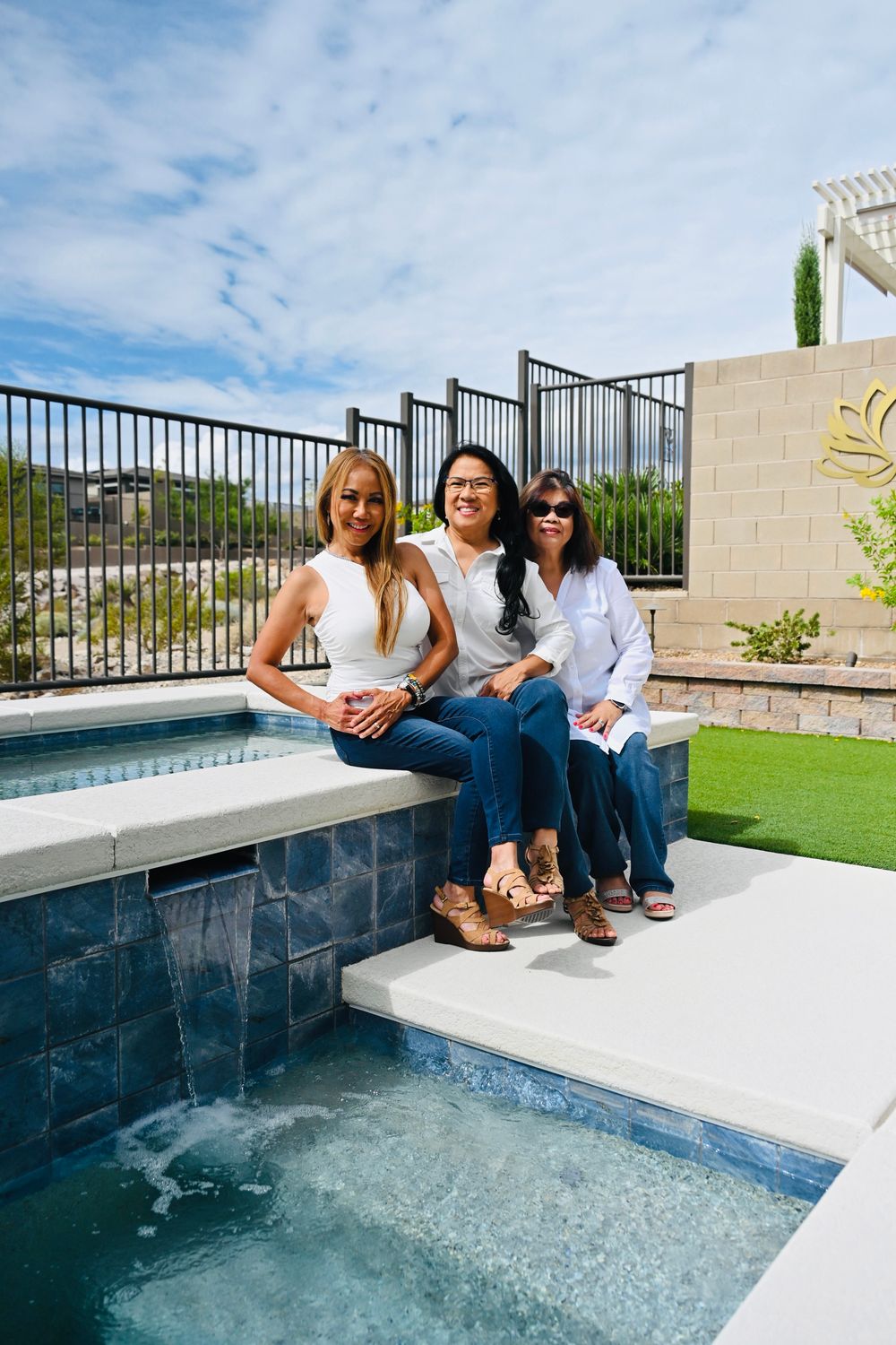 Three women in white tops and jeans sitting together by outdoor pool.