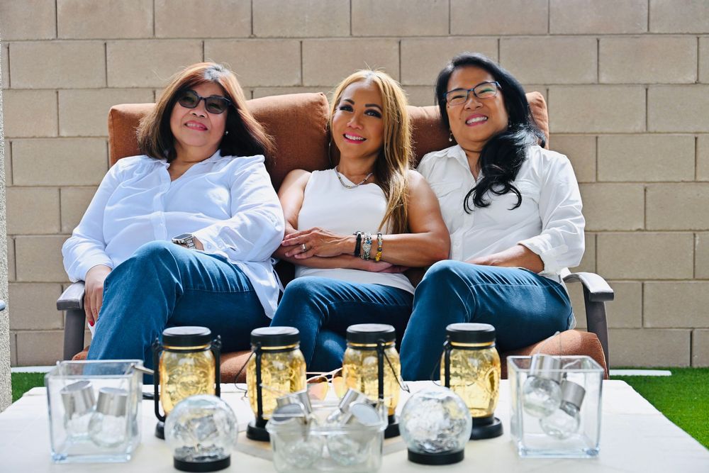Three smiling women wearing white shirts and jeans sit together on a patio sofa with decorative mason jar lights displayed.