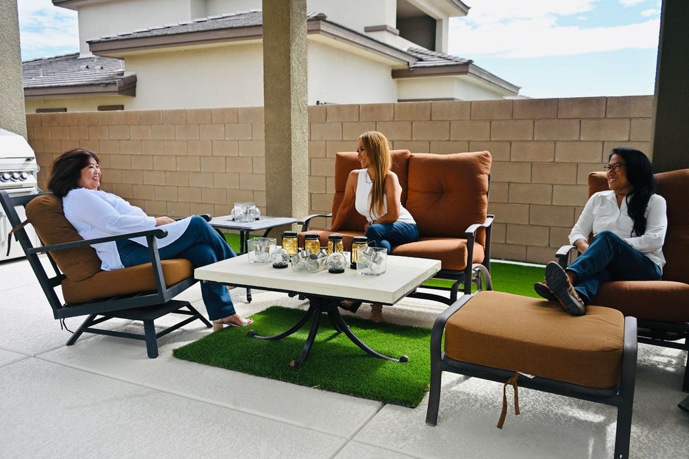 Three women in white tops enjoy conversation while seated on outdoor patio furniture with drinks on a white coffee table.