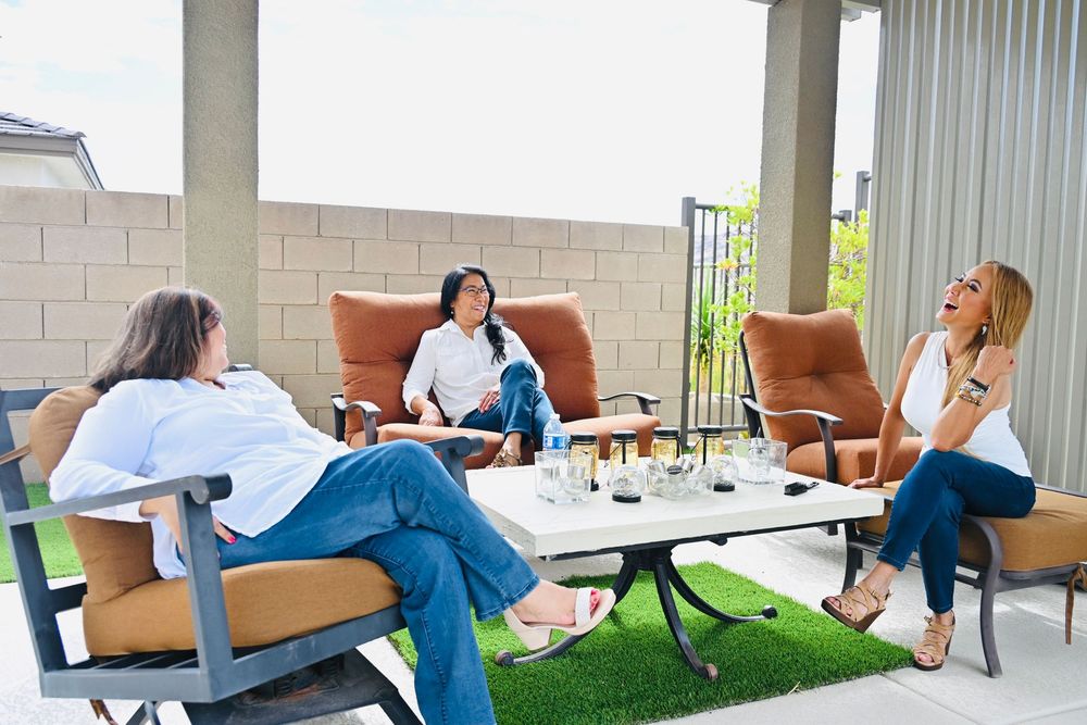 Three women in casual white outfits laugh and chat together on an outdoor patio with artificial grass and mountain views.