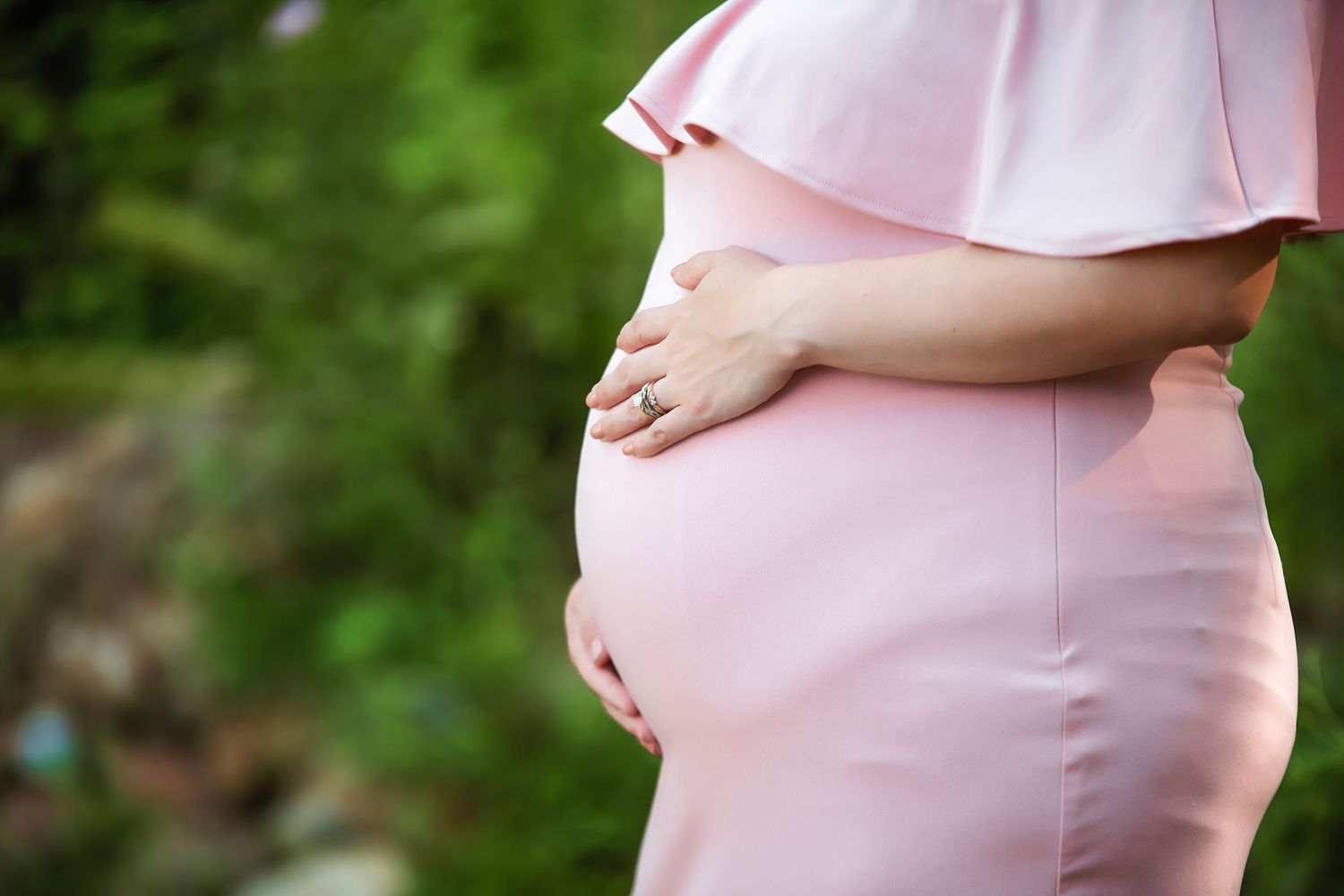 Pregnant woman in pink dress cradles her baby bump against a green outdoor background.