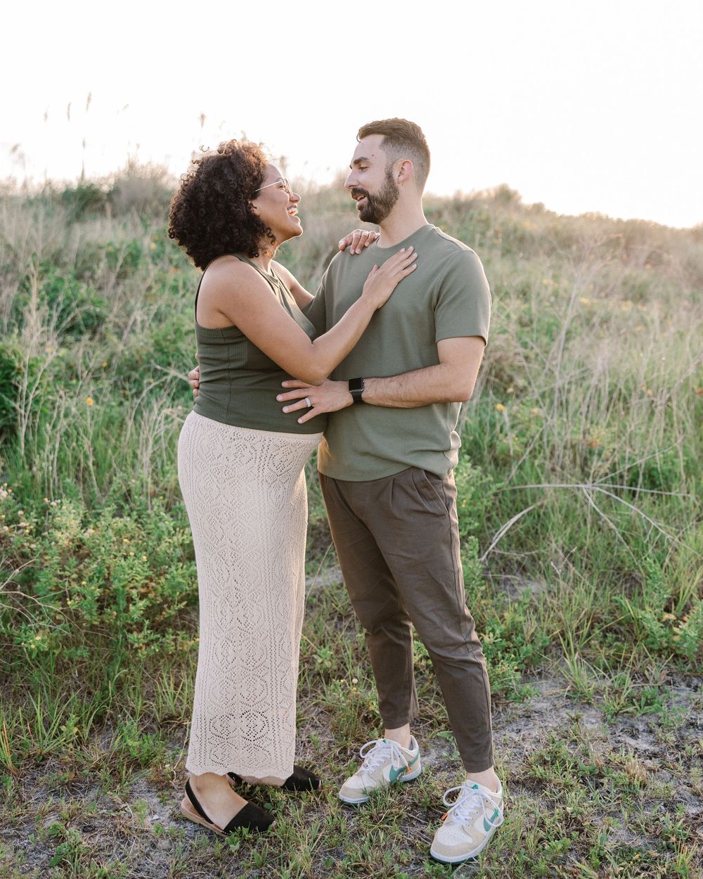 A couple in coordinated olive green outfits share intimate moments in a grassy meadow at sunset.