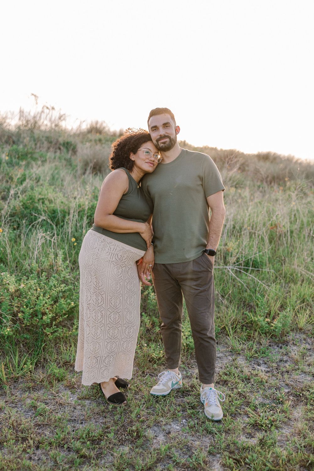 A couple embraces while standing in a grassy field at sunset, wearing casual outfits in earth tones.