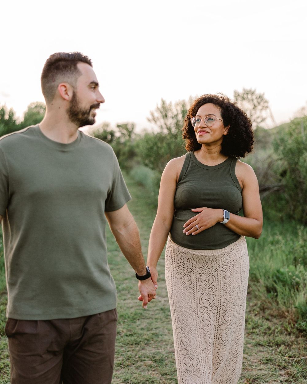 Romantic couple in casual attire embrace while standing in a sunlit field with natural greenery.